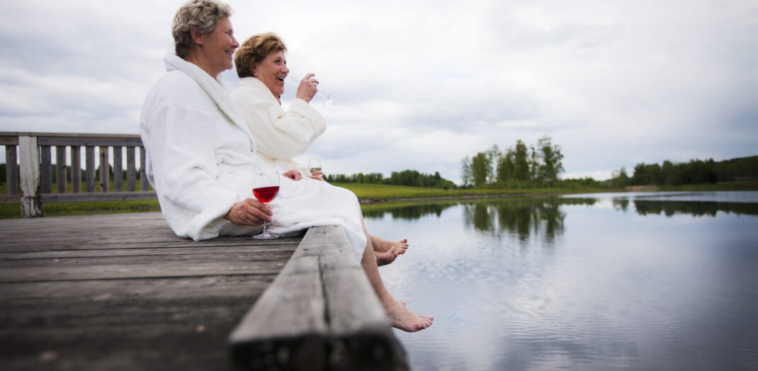 Två kvinnor i vita morgonrockar som dricker rödvin på en brygga.
