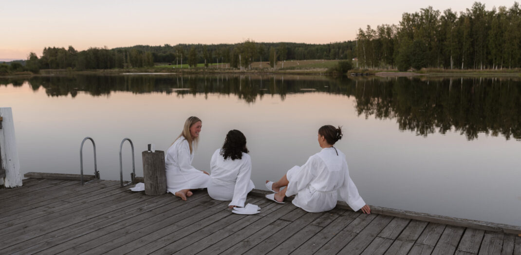 3 kvinnor i vit badrock sitter på bryggan vid sjön Fryken i Sunne