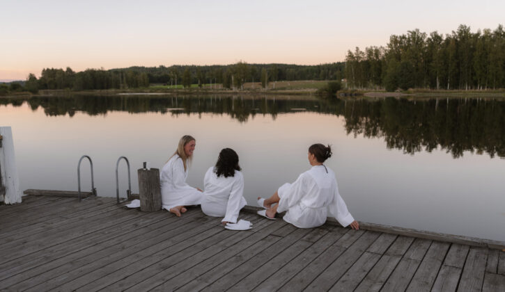 3 kvinnor i vit badrock sitter på bryggan vid sjön Fryken i Sunne